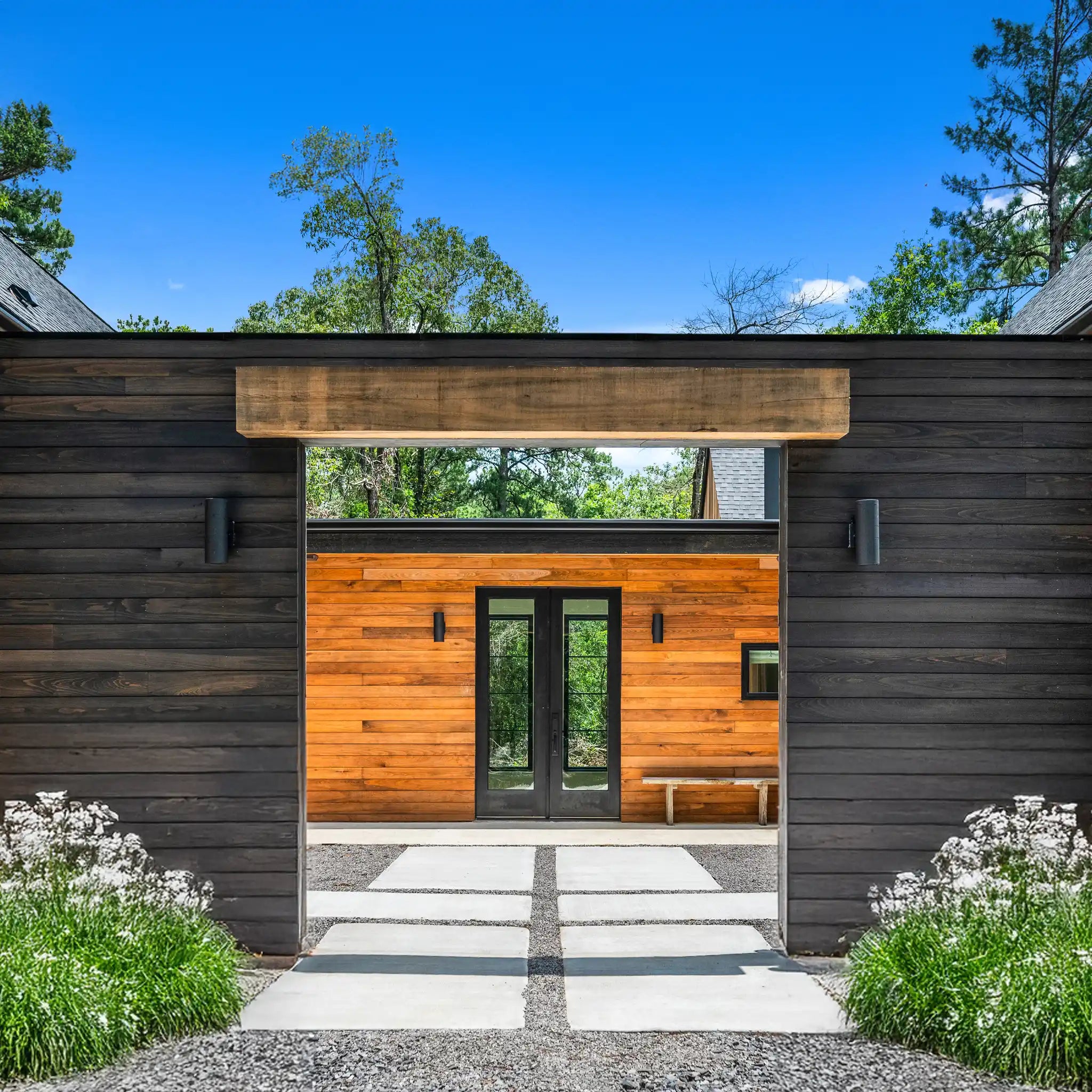 Modern farmhouse entry courtyard with dark wood siding, a warm wood-paneled entryway, double glass doors, a stone walkway, and landscaped greenery under a blue sky.