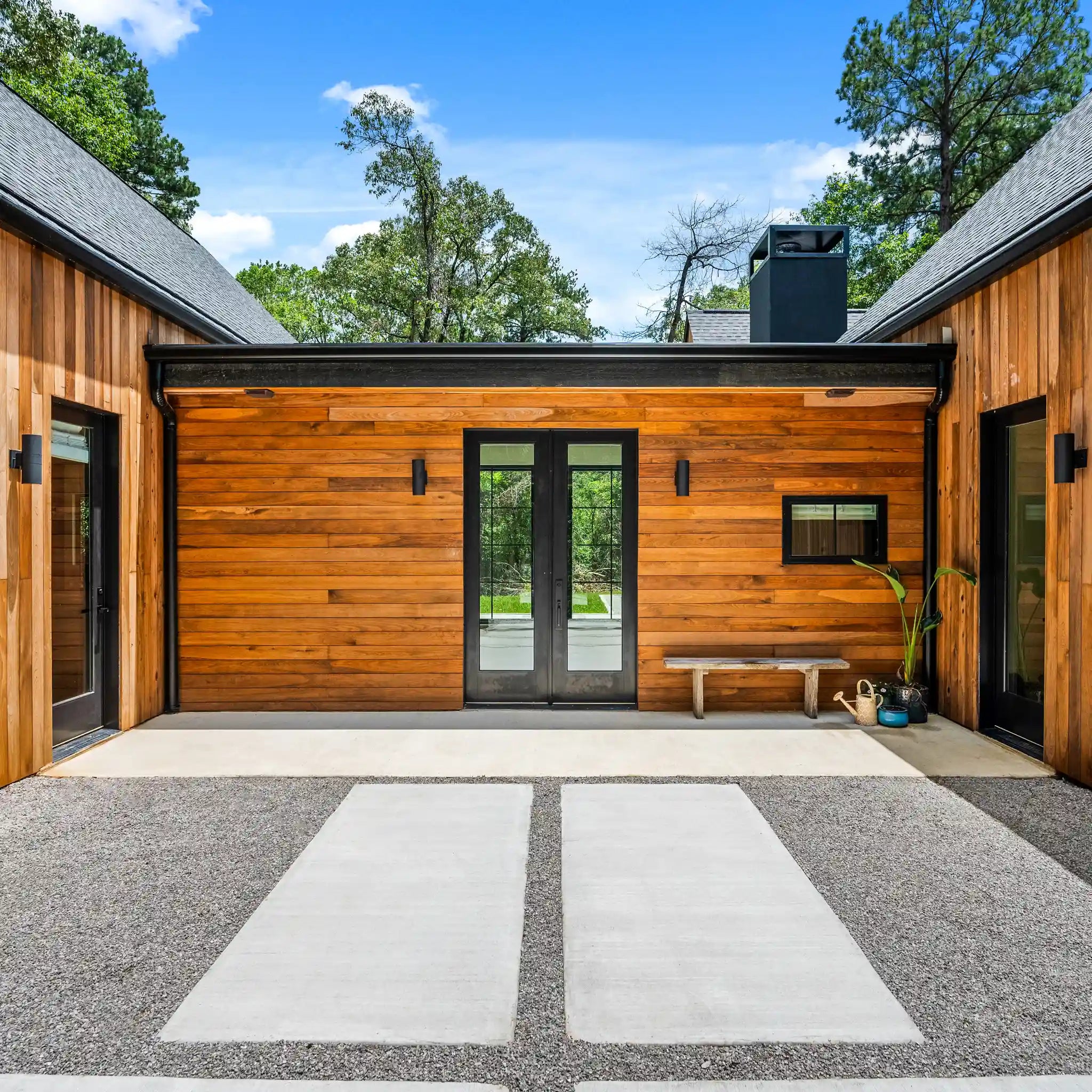 Modern farmhouse exterior entry courtyard with a gravel path, concrete pavers, black-framed double doors, and a small bench against the wall under a blue sky with trees.