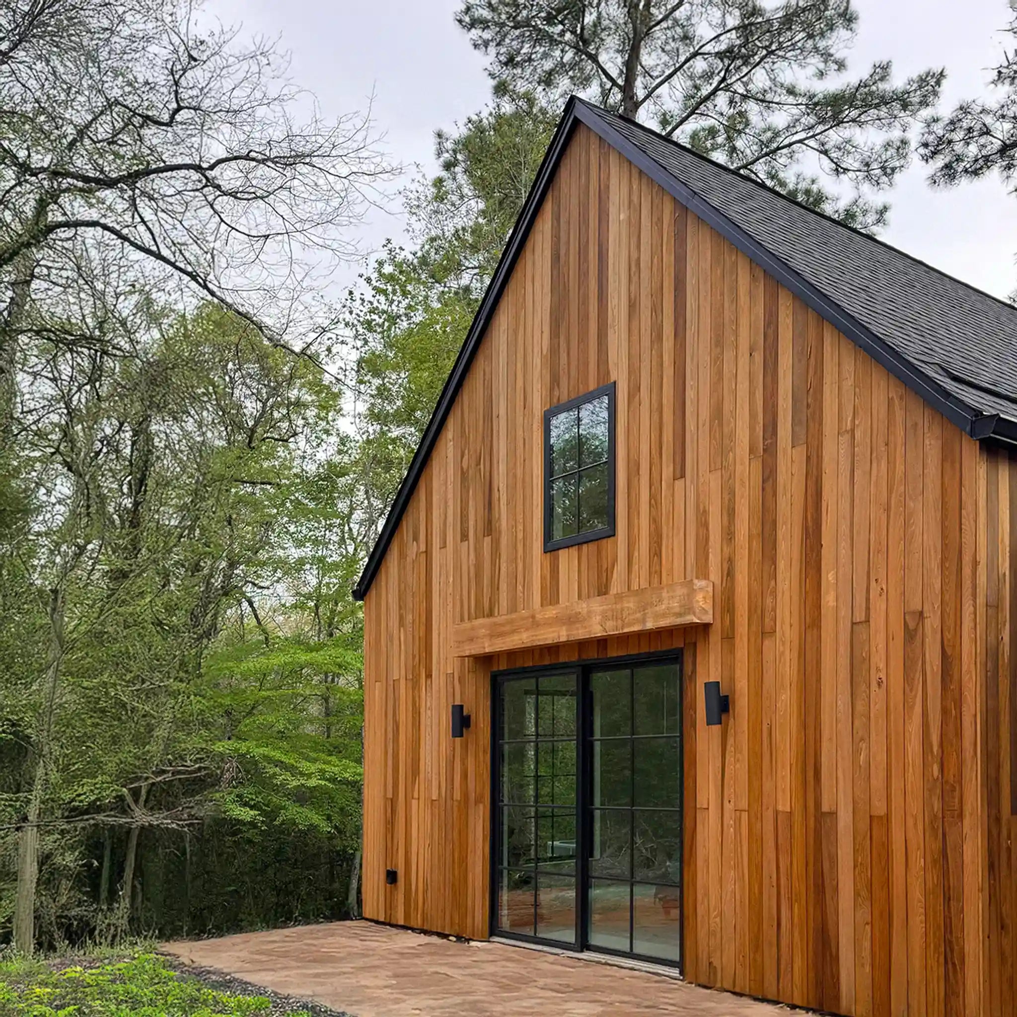 Modern wooden cabin exterior with warm medium-brown vertical wood siding, black metal roof, black-framed windows, and a paved entryway surrounded by green trees.