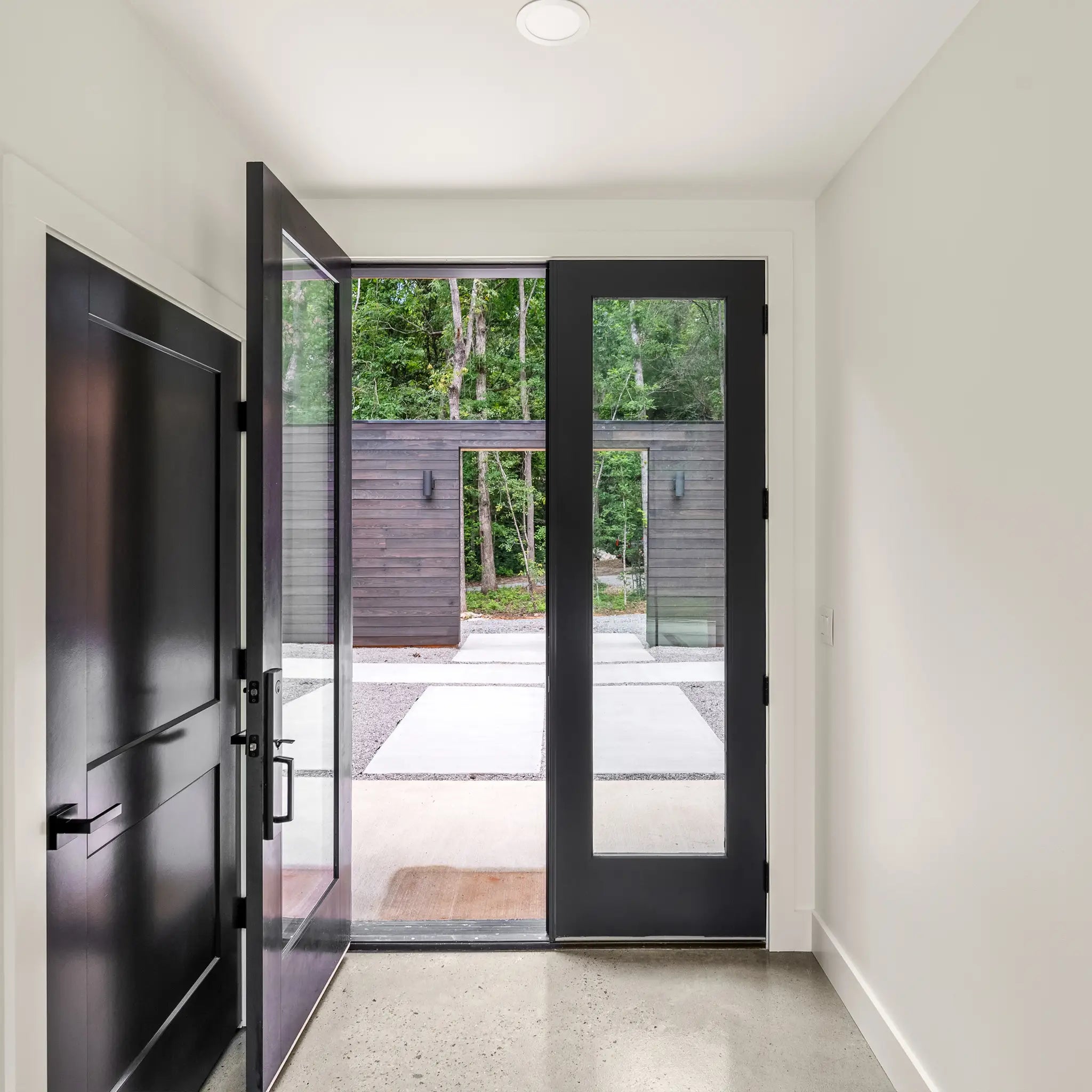 Modern black metal double glass doors with an open left door, showing a concrete patio and outdoor greenery beyond, set within a clean white-walled entryway.