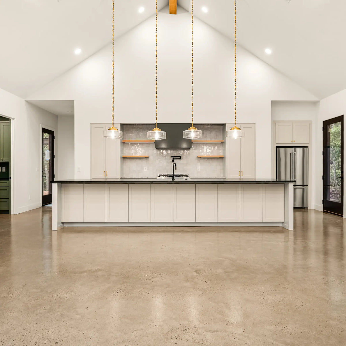 A modern farmhouse kitchen island with white cabinets and a black countertop, set in a bright home with concrete floors and three hanging pendant lights.