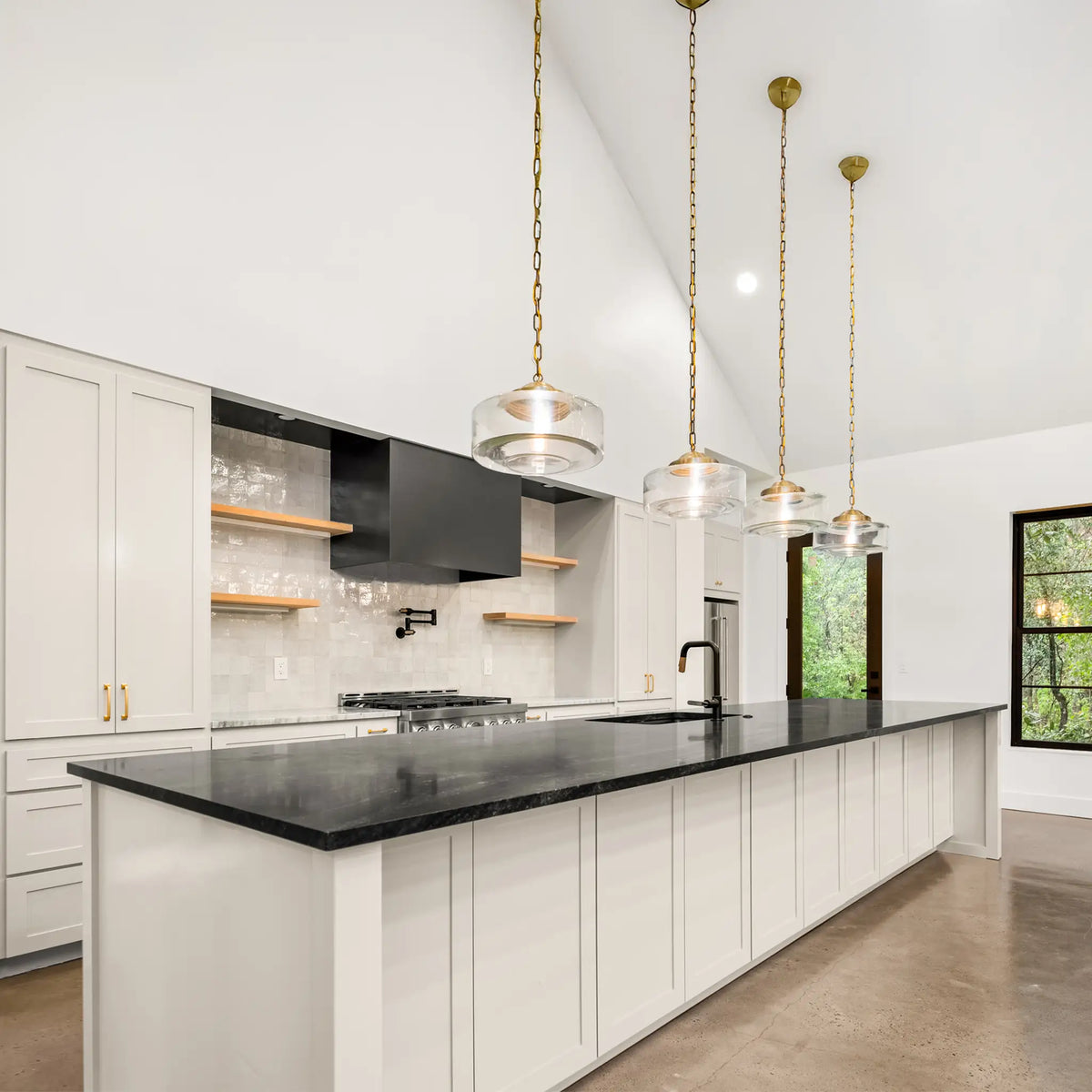 A modern kitchen island with a black countertop, white cabinetry, and three gold pendant lights hanging above. The kitchen features white cabinets, a black range hood, and a light stone backsplash.