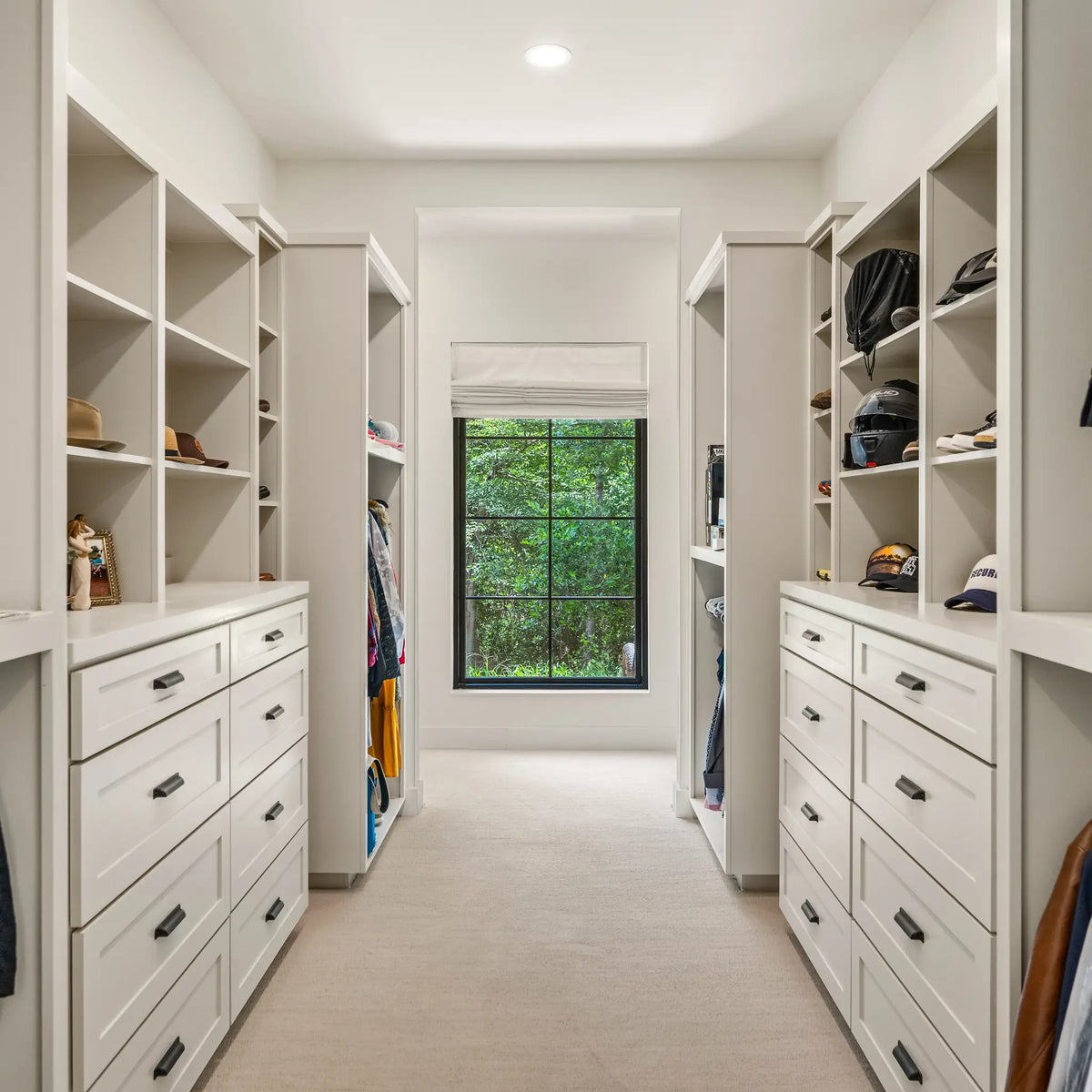 A bright, modern walk-in closet with white shelving and drawers on both sides, a window at the back showing green trees outside, and neutral-toned clothing and accessories displayed on shelves.
