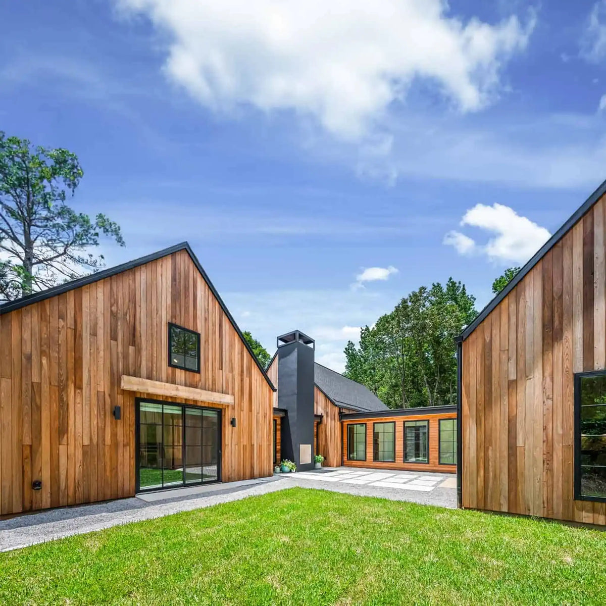 Modern wooden cabin exterior with warm wood siding, large glass windows, a central chimney, and a grassy courtyard under a blue sky with clouds.