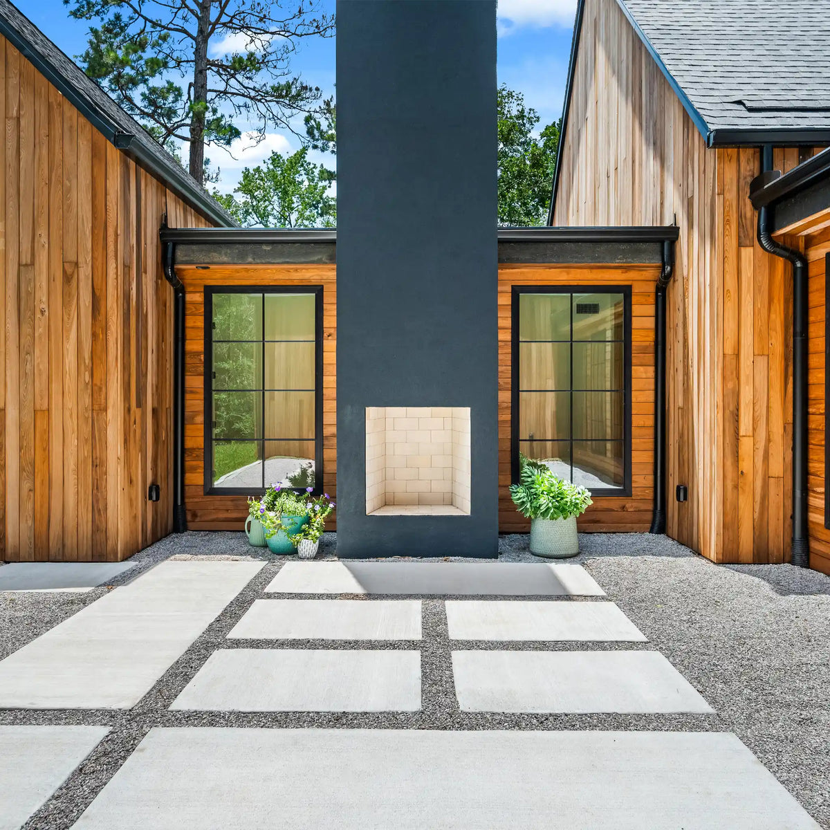 Modern minimalist farmhouse outdoor patio area with a central dark gray chimney, a built-in stone fireplace niche, wood-clad buildings, gravel ground cover, and large light stone pavers.